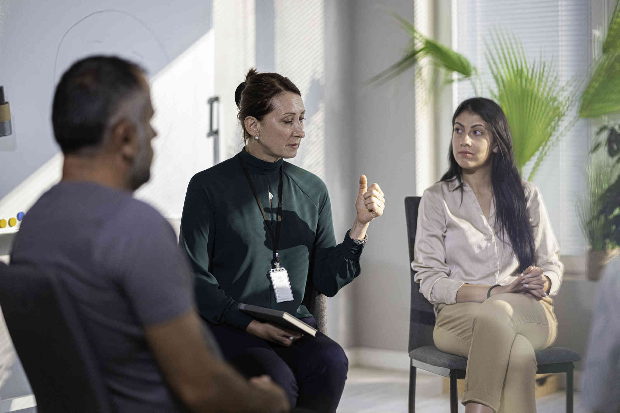 A woman in a green blouse wearing an ID badge around her neck speaks while a man and a woman sitting next to her listen.